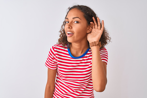 Young brazilian woman wearing red striped t-shirt standing over isolated white background smiling with hand over ear listening an hearing to rumor or gossip. Deafness concept.