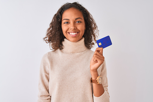 Young brazilian woman holding credit card standing over isolated white background with a happy face standing and smiling with a confident smile showing teeth