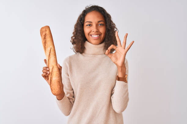 Young brazilian baker woman holding bread standing over isolated white background doing ok sign with fingers, excellent symbol