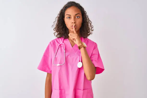 Photo of Young brazilian nurse woman wearing stethoscope standing over isolated white background asking to be quiet with finger on lips. Silence and secret concept.