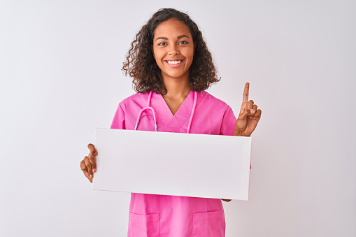 Young brazilian nurse woman holding banner standing over isolated white background surprised with an idea or question pointing finger with happy face, number one