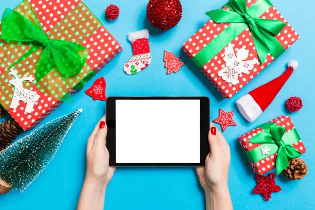 Photo of Top view of woman holding tablet in her hands on blue background made of Christmas decorations. New Year holiday concept. Mockup