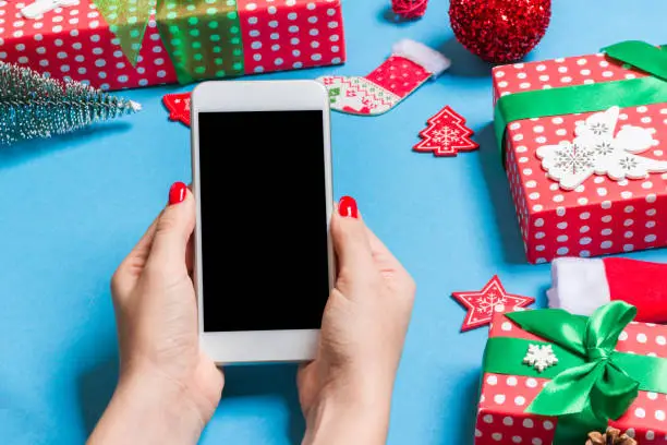 Photo of Top view of phone in female hand on festive blue background. Christmas decorations. New Year time holiday. Mockup