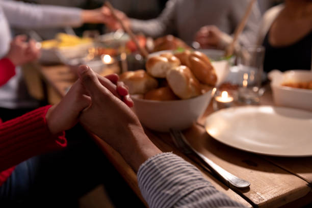 Millennial adult friends celebrating Thanksgiving together at home Front view mid section of a group of young adult multi-ethnic male and female friends sitting around a table holding hands saying grace before eating Thanksgiving dinner at home together saying grace stock pictures, royalty-free photos & images