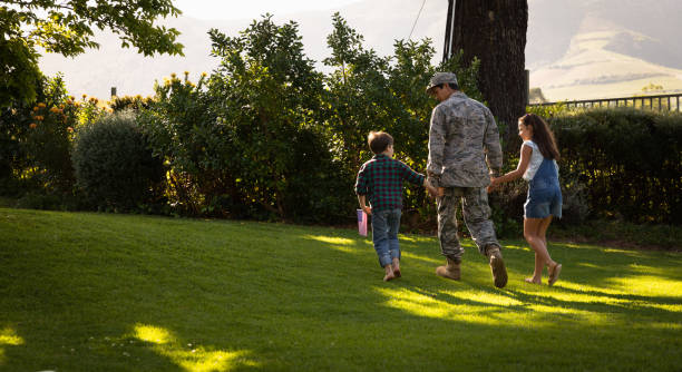 Rear view of a young adult mixed race male soldier in the garden outside his home, holding hands and walking with his young son and daughter, his son holding a US flag