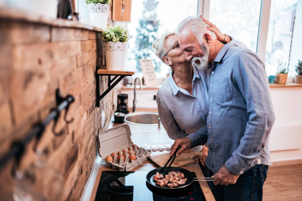 couples aînés affectueux préparant la nourriture à la maison. la femme embrasse son homme. - cooking senior adult healthy lifestyle couple photos et images de collection