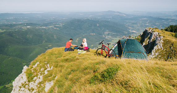 Side view of couple sitting at edge of cliff while camping in mountains and cooking.