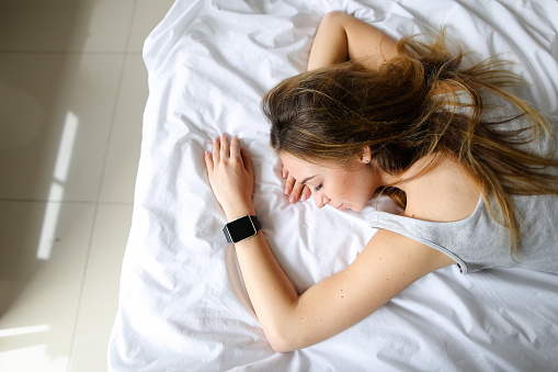 Young american woman lying on bed with smart watch, white linens background. Concept of modern gadgets and technology.