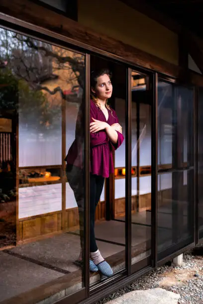 Young cold shivering woman standing in Japan looking through open window sliding door on indoor Japanese traditional garden with gravel stone rocks in house