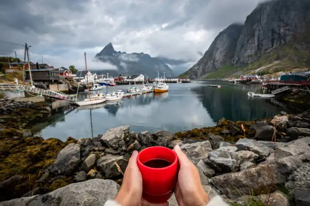 POV photo of a woman having a coffee in the fishing village of Hamnøy in the Lofoten Islands with mountain range and cloudy sky in background during summer.