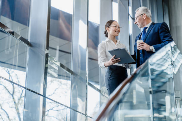 senior geschäftsmann und junge asiatische frau arbeiten mit dokumenten zusammen in büro-lobby - business two people talking building exterior stock-fotos und bilder