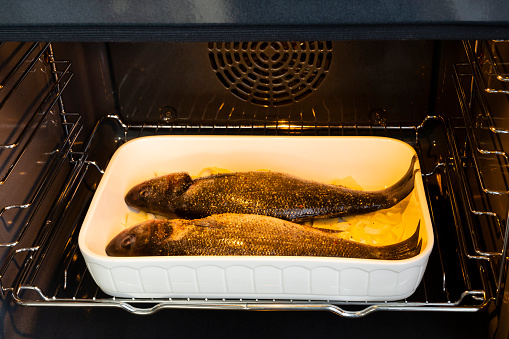 a white ceramic baking dish with two raw seabass fishes on a layer of cut onion are in an oven before being baked. Close up view.