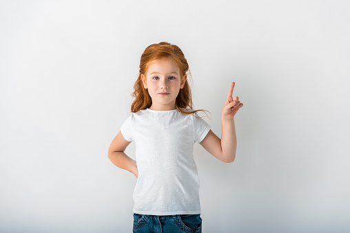 cute redhead kid looking at camera and pointing with finger on white