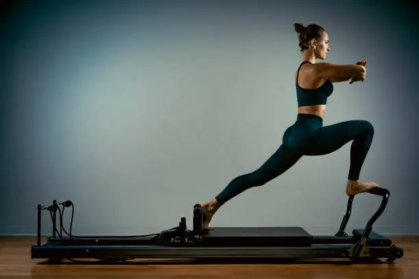 Young girl doing pilates exercises with a reformer bed. Beautiful slim fitness trainer on a reformer gray background, low key, art light, copy space advertising banner.