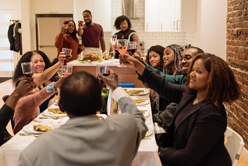 Family time for Thanksgiving dinner. Multi-generation members of african-american family sitting at the table, chatting and cheering. Modern appartement with brick wall. Horizontal indoors waist up shot with some copy space.