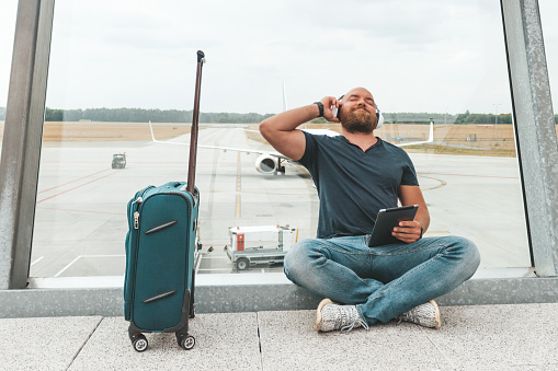 Bearded hipster traveller is waiting for his flight and listening music in front of the airfield