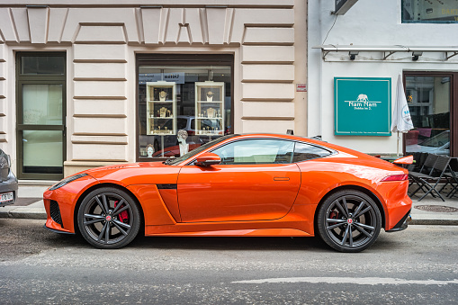 An orange colored Jaguar F-Type SVR coupe is parked on a street in Vienna Austria on a cloudy day.
