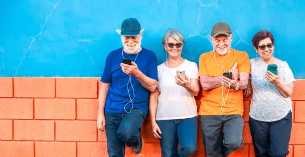 Photo of Two happy couples of senior people standing against an orange and blue wall smiling and looking at the mobile phone using earphones. Casual clothing and relaxed faces