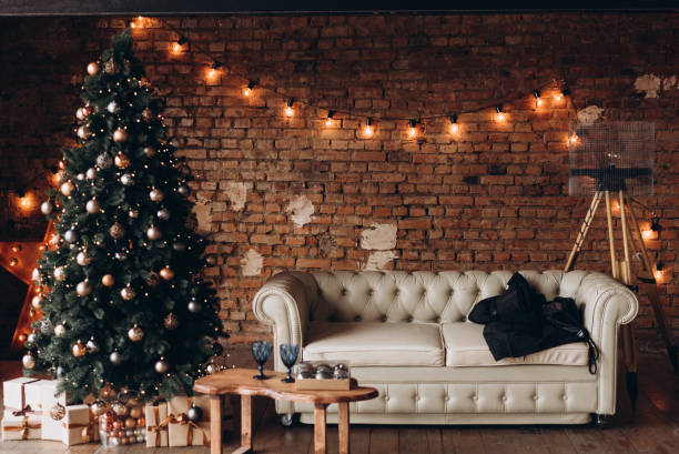 hermoso árbol de navidad decorado con juguetes y guirnaldas cerca de un sofá de cuero blanco contra una pared de ladrillo - fashionable party design home decorating fotografías e imágenes de stock