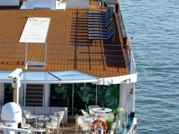 cruise boat upper deck detail in bright summer light with inviting brown flooring and a row of lounge chairs. white tables, chairs at the restaurant level. green plants. orange floating redcue device