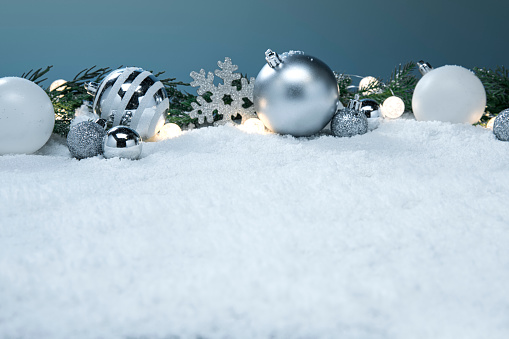 This is a photograph of blue and white and Silver Christmas ornaments shot in the snow surrounded by glowing Christmas lights surrouned by evergreen palm tree branches. There are no people in the photograph