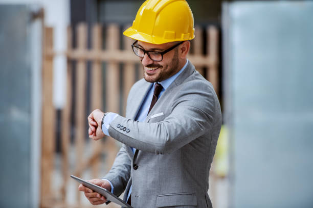 スーツを着た魅力的なエレガントな白人の笑顔の建築家で、メガネとヘルメットの頭が建設現場に立ち、腕時計を見ながらタブレットを持っています。 - construction site inspection interview ストックフォトと画像
