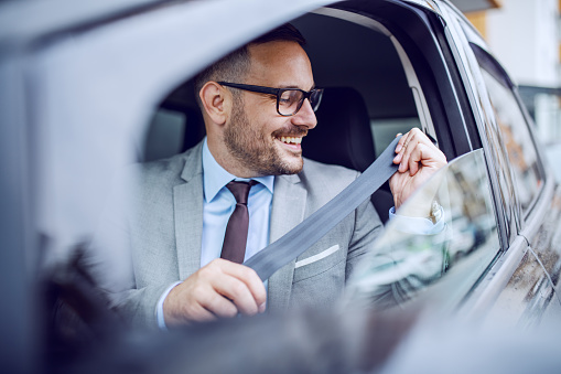 Attractive classy Caucasian smiling businessman in suit and with eyeglasses buckling up safety belt in his car.