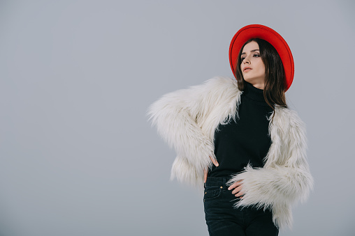 attractive elegant woman posing in white fur coat and red hat, isolated on grey