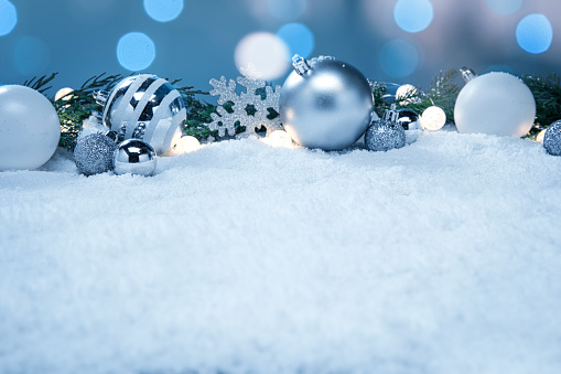 This is a photograph of blue and white and Silver Christmas ornaments shot in the snow surrounded by glowing Christmas lights surrouned by evergreen palm tree branches. There are no people in the photograph