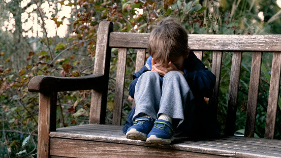 Crying caucasian child boy sitting on a schoolyard bench covering his face