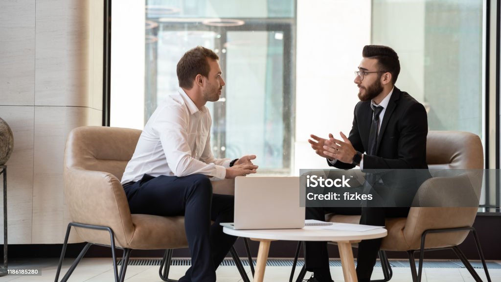Middle eastern and caucasian ethnicity businessmen talking negotiating indoors Middle eastern and caucasian ethnicity businessmen seated on armchair in modern office talking solve common issues, banker telling to client regarding bank services make recommendations and consulting Discussion Stock Photo