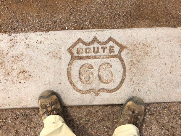 Route 66 Logo Engraved in Pavement Historic US Highway 66, known as the “Mother Road”, is more than just a stretch of pavement. It is also an American icon, a symbol of opportunity, adventure and discovery. US Highway 66 better known in literature, song, and story as Route 66, was a ribbon of roadway over two thousand miles long that connected Middle America to the Pacific coast. Beginning in 1926, Route 66 fulfilled different needs in each subsequent decade. In the 1930’s it was the main travel corridor for migrating families fleeing the Dust Bowl. In the 1940’s, during World War II, the road was filled with military traffic. In the 1950’s Route 66 came into its own as motorists took to the road to explore the nation with a freedom never felt before. In the next decades as the interstate highway system was being developed, drivers bypassed Route 66 in favor of the faster freeways. Unfortunately, the tourists also bypassed the small towns that gave the Mother Road its character and appeal. Petrified Forest is the only National Park in the country that contains a section of Historic Route 66. This stretch of Route 66 was open from 1926 until 1958 and was the primary way millions of travelers accessed the Petrified Forest and Painted Desert. Near Tiponi Point and the Painted Desert a section of the original roadbed has been preserved along with interpretive signs and a rusty 1932 Studebaker. The Route 66 logo is engraved in the pavement at the Route 66 interpretive center in Petrified Forest National Park near Holbrook, Arizona, USA. jeff goulden petrified forest national park stock pictures, royalty-free photos & images