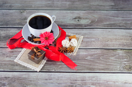 A cup of black coffee on a saucer decorated with spices, flowers and a scarlet ribbon, next to a napkin with a gift box. Close-up.