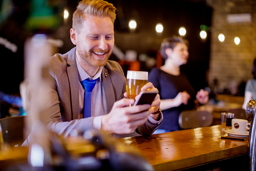 Smiling young businessman sitting at the bar counter with pint of craft beer and mobile phone