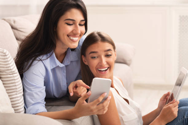 istock Two Girls Using Phone And Tablet Sitting On Sofa Indoor 1186253636