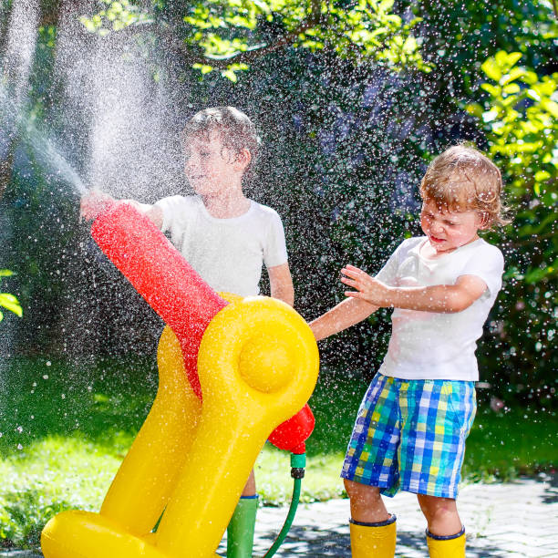 Two little kids boys playing with a garden hose sprinkler on hot and sunny summer day. Children having fun with sprinkling water, drops rain. Outdoors leisure wth water for friends. Rain rubber boots dos niños pequeños jugando con un aspersor de manguera de jardín en el caluroso y soleado día de verano. los niños divirtiéndose con agua espolvoreada, deja caer la lluvia. al aire libre ocio wth agua para los amigos. botas de goma de lluvia.