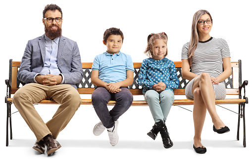 Full length portrait of a mother, father and children sitting on a bench and looking at the camera isolated on white background