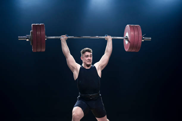 Male weightlifter performing clean and jerk lift Young male weightlifter lifting barbell above his head while performing clean and jerk lift. clean and jerk stock pictures, royalty-free photos & images