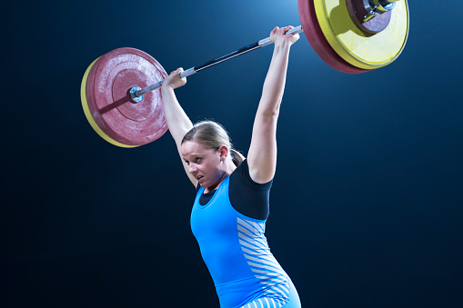 Young female weightlifter lifting barbell above her head while performing clean and jerk lift.