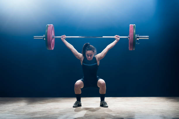 Female weightlifter performing clean and jerk lift Young female weightlifter lifting barbell above her head while performing clean and jerk lift. clean and jerk stock pictures, royalty-free photos & images