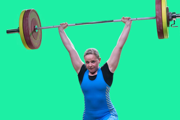 Female weightlifter performing clean and jerk lift Young female weightlifter lifting barbell above her head while performing clean and jerk lift. clean and jerk stock pictures, royalty-free photos & images