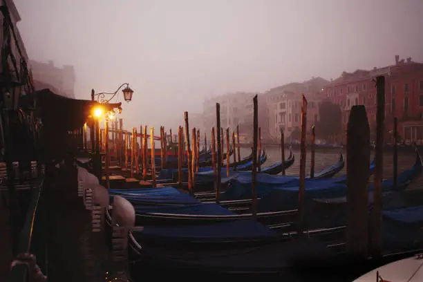 Venice, Italy - October 28, 2019: empty gondolas are parked to the poles on the Grand Canal in the dark early morning mist just before the sunrise, no people are visible, street lamps illuminate calm river and beautiful old boats in the historical town.