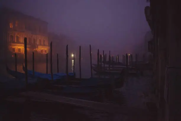 Venice, Italy - October 28, 2019: empty gondolas are parked to the poles on the Grand Canal in the dark early morning mist just before the sunrise, no people are visible, street lamps illuminate calm river and beautiful old boats in the historical town.