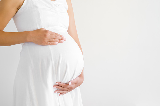 Pregnant woman in white dress touching big belly with hands. Baby expectation. Pregnancy time - 37 weeks. Closeup. Empty place for text on gray background.