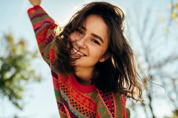 Bottom view close-up portrait of happy young woman smiling broadly with windy hair and freckles has joyful expression, wearing colorful knitted sweater with arms wide open, posing on nature sunlight stock photo