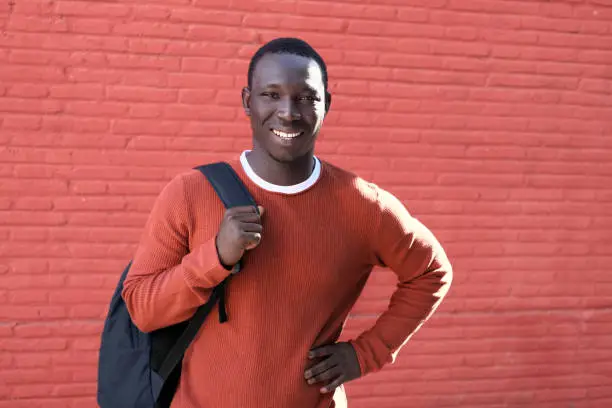 Portrait Of African Young Man Looking At Camera And Smiling, Carrying Backpack For School. Copy Space On Red Wall In Background.