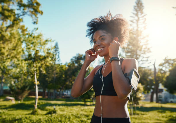 lycklig ung svart afrikansk amerikansk kvinna njuter av att lyssna på musik på hörlurar medan jogging träning i det ljusa solljus på park - park run bildbanksfoton och bilder