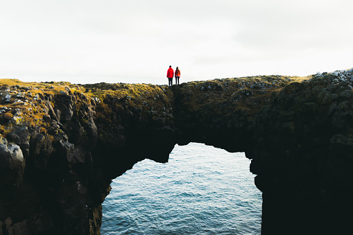 Young woman and man in red jackets enjoying their journey at Snaefellsnes Peninsula in West Iceland, walking at huge natural arch above the sea