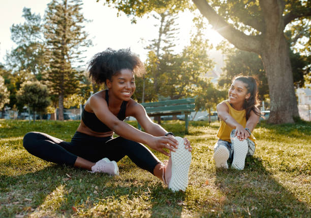 jeunes amis féminins divers s'asseyant sur l'herbe verte étirant ses jambes dans la lumière du soleil de matin au stationnement - amis divers se réchauffant avant de faire l'exercice de groupe - échauffement photos et images de collection