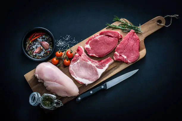 Raw meat assortment - fillet steak, chicken breast and pork chops shot from above on dark background. The cuts of meat are on a wooden cutting board and some ingredients and spices like peppercorns, rosemary, marine salt, garlic and tomatoes are all around them. Predominant colors are red and black. XXXL 42Mp studio photo taken with Sony A7rii and Sony FE 90mm f2.8 macro G OSS lens